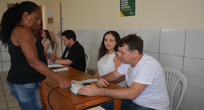 Em uma sala simples, com paredes claras e azulejos, uma mesa de madeira está posicionada no cent...