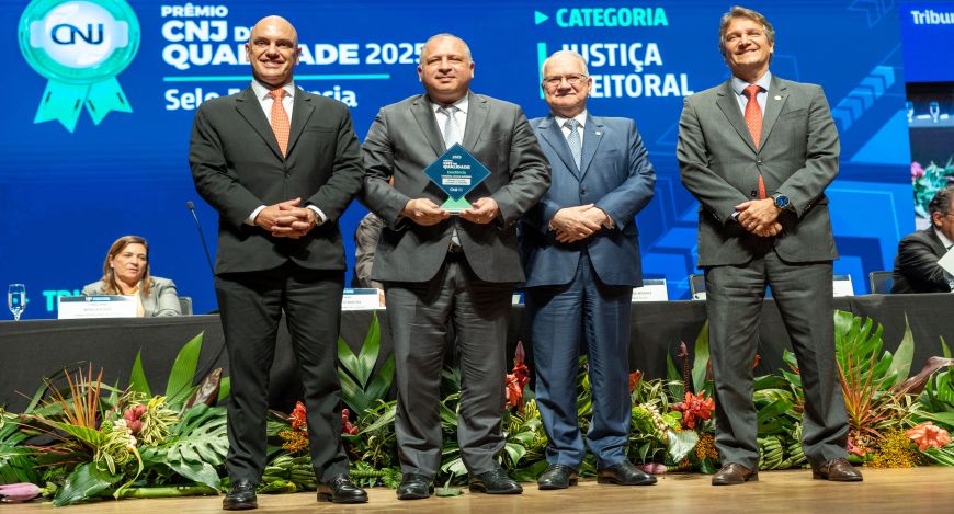 TRE de Roraima é reconhecido como o melhor tribunal eleitoral do país A imagem mostra uma cerimônia de premiação em um palco. Quatro pessoas estão em pé na frente, ve...