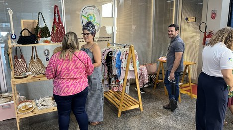Ambiente interno com feira de artesanato. Há vários expositores com bolsas, roupas infantis e ac...