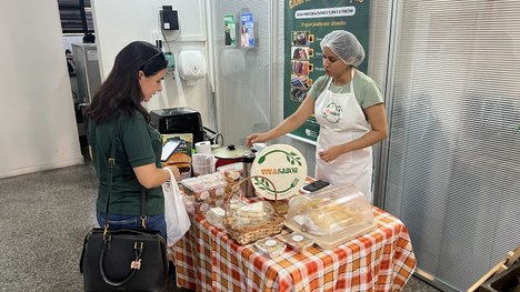 Ambiente interno com feira de artesanato. Há vários expositores com bolsas, roupas infantis e ac...