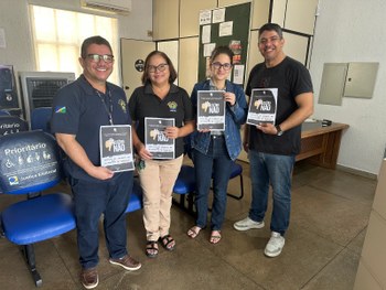 Quatro adultos estão em pé, lado a lado, em um ambiente interno de atendimento público. São dois...