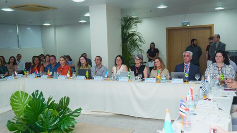 A foto mostra um grupo de pessoas sentadas ao redor de mesas em formato de “U”, participando de ...