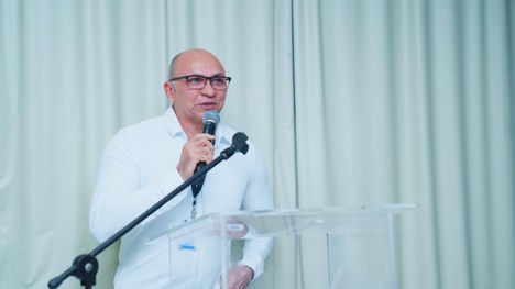 A foto mostra um grupo de pessoas sentadas ao redor de mesas em formato de “U”, participando de ...