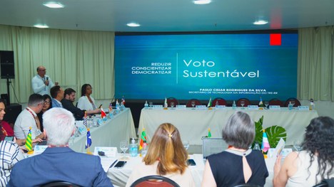 A foto mostra um grupo de pessoas sentadas ao redor de mesas em formato de “U”, participando de ...