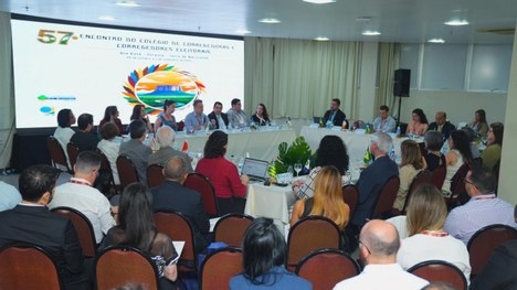 A foto mostra um grupo de pessoas sentadas ao redor de mesas em formato de “U”, participando de ...