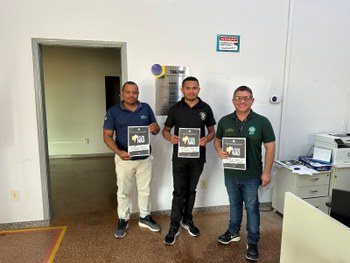 Quatro adultos estão em pé, lado a lado, em um ambiente interno de atendimento público. São dois...