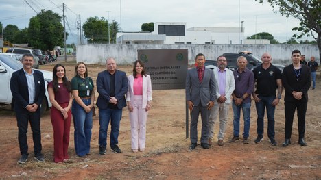 Grupo de cerca de doze adultos posa em pé ao ar livre, em um terreno de terra batida. Eles estão...
