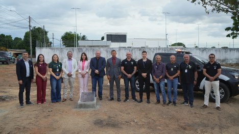 Grupo de cerca de doze adultos posa em pé ao ar livre, em um terreno de terra batida. Eles estão...