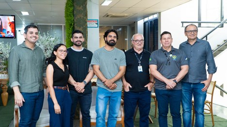 A imagem mostra oito pessoas em pé, alinhadas lado a lado em um ambiente interno que parece ser ...