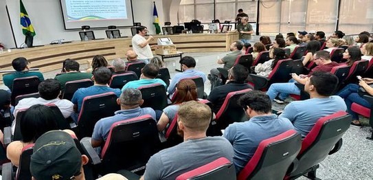 Foto de uma capacitação em auditório. Participantes estão sentados em fileiras acompanhando uma ...