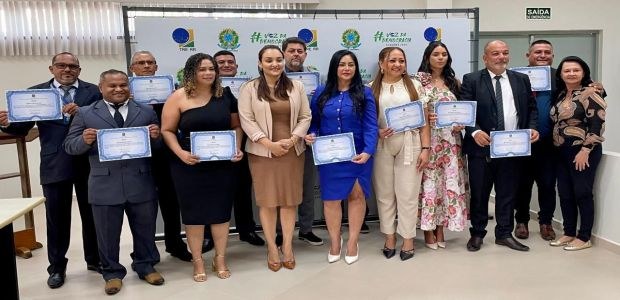 Diplomação Caracarai Em uma sala de paredes brancas, treze pessoas adultas, entre homens e mulheres, estão em pé, à f...