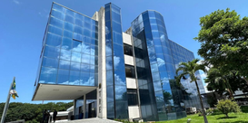 Prédio moderno de vários andares, com fachada de vidro azul espelhado que reflete o céu e nuvens...