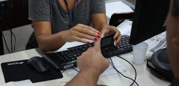 Foto do momento da coleta das digitais de um eleitor, a imagem mostra apenas as mãos das pessoas...