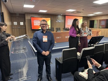 A imagem mostra um auditório cheio, com várias pessoas assistindo a três palestrantes sentados à...