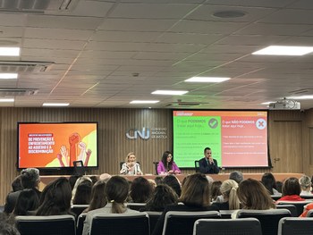 A imagem mostra um auditório cheio, com várias pessoas assistindo a três palestrantes sentados à...