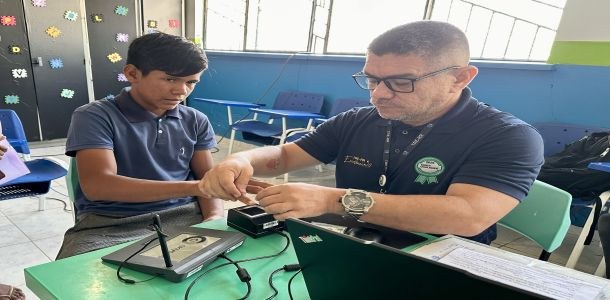 Equipes da Justiça Eleitoral Itinerante atendem em Rorainópolis e Pacaraima até esta sexta-feira, 13 A imagem mostra uma cena em uma sala com carteiras escolares ao fundo. Uma pessoa está sentada à...