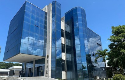 A imagem mostra um prédio de vários andares, com uma fachada moderna feita principalmente de vid...