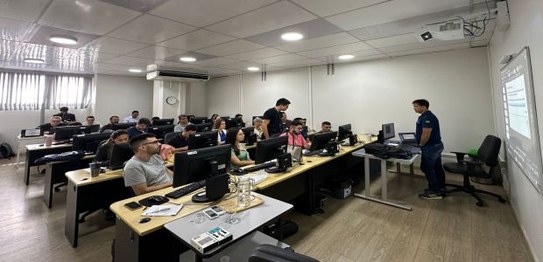 Na foto várias pessoas entre homens e mulheres estão em uma sala de paredes brancas e mesas com ...