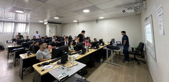 Na foto várias pessoas entre homens e mulheres estão em uma sala de paredes brancas e mesas com ...