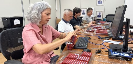 Quatro pessoas sentadas lado a lado em uma sala de trabalho, manuseando e organizando pequenos d...