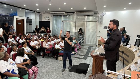 Um auditório está cheio de pessoas idosas sentadas em cadeiras vermelhas, muitas usando camiseta...