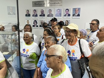 Um auditório está cheio de pessoas idosas sentadas em cadeiras vermelhas, muitas usando camiseta...