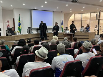 Um auditório está cheio de pessoas idosas sentadas em cadeiras vermelhas, muitas usando camiseta...