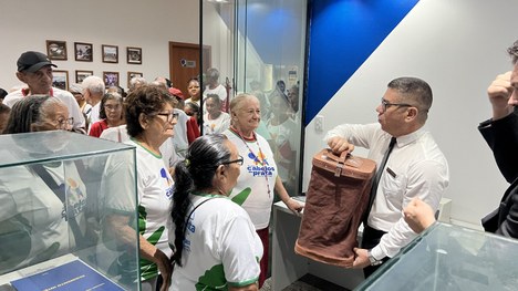 Um auditório está cheio de pessoas idosas sentadas em cadeiras vermelhas, muitas usando camiseta...