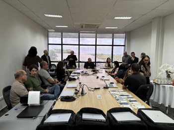 Foto de uma mesa em ambiente interno com várias mochilas pretas alinhadas. Sobre cada mochila há...