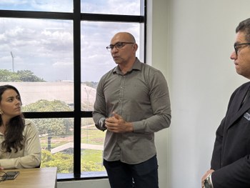 Foto de uma mesa em ambiente interno com várias mochilas pretas alinhadas. Sobre cada mochila há...