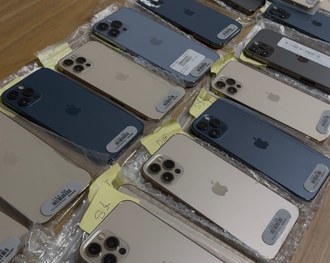 Foto de uma mesa em ambiente interno com várias mochilas pretas alinhadas. Sobre cada mochila há...