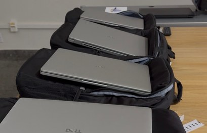 Foto de uma mesa em ambiente interno com várias mochilas pretas alinhadas. Sobre cada mochila há...