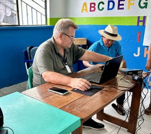 Em uma sala com paredes coloridas em azul, verde e branco, uma pessoa está sentada à mesa operan...
