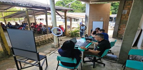 A foto mostra um espaço simples, parecido com uma área coberta de uma comunidade rural. No prime...