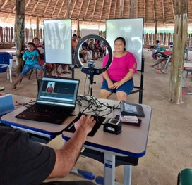 A imagem mostra uma mulher sentada diante de uma câmera com aro de luz, enquanto um servidor ope...