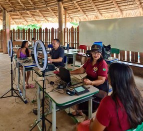 A imagem mostra uma mulher sentada diante de uma câmera com aro de luz, enquanto um servidor ope...