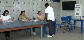 Quatro pessoas em um local de votação, três sentadas atrás de uma mesa e uma em pé sendo atendid...