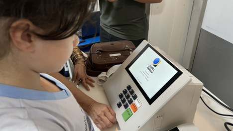 A imagem mostra uma pessoa posicionada diante de uma urna eletrônica. A mão dessa pessoa está ap...