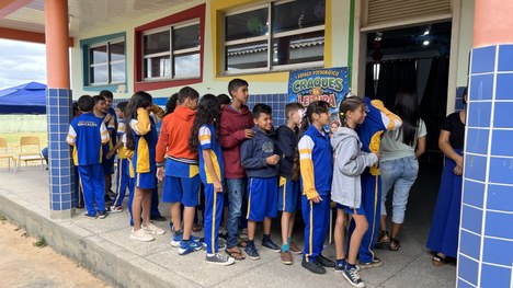 A imagem mostra quatro crianças usando uniformes escolares claros sentadas próximas a uma mesa c...