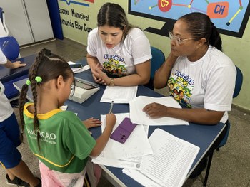 A imagem mostra quatro crianças usando uniformes escolares claros sentadas próximas a uma mesa c...