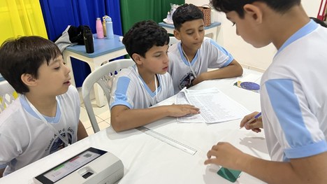 A imagem mostra quatro crianças usando uniformes escolares claros sentadas próximas a uma mesa c...