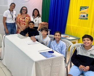 A imagem mostra quatro crianças usando uniformes escolares claros sentadas próximas a uma mesa c...