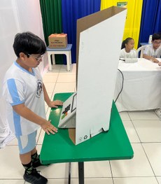A imagem mostra quatro crianças usando uniformes escolares claros sentadas próximas a uma mesa c...