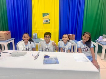 A imagem mostra quatro crianças usando uniformes escolares claros sentadas próximas a uma mesa c...