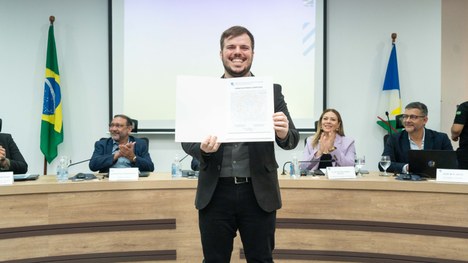 A imagem mostra um auditório cheia de pessoas em pé. As cadeiras são vermelhas e estão organizad...