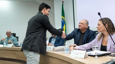 A imagem mostra um auditório cheia de pessoas em pé. As cadeiras são vermelhas e estão organizad...