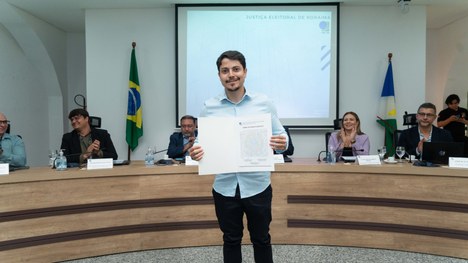 A imagem mostra um auditório cheia de pessoas em pé. As cadeiras são vermelhas e estão organizad...