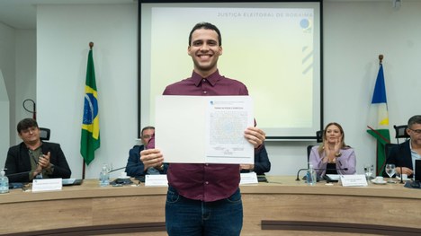 A imagem mostra um auditório cheia de pessoas em pé. As cadeiras são vermelhas e estão organizad...