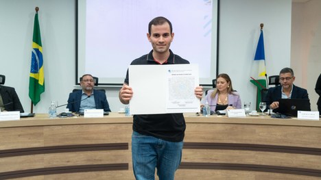 A imagem mostra um auditório cheia de pessoas em pé. As cadeiras são vermelhas e estão organizad...