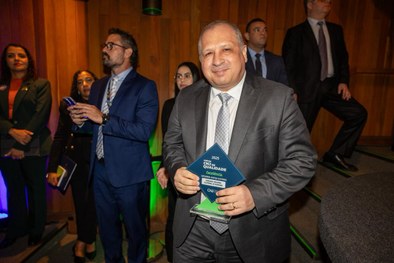 A imagem mostra um homem de terno cinza claro sorrindo e segurando um troféu azul do Prêmio CNJ ...